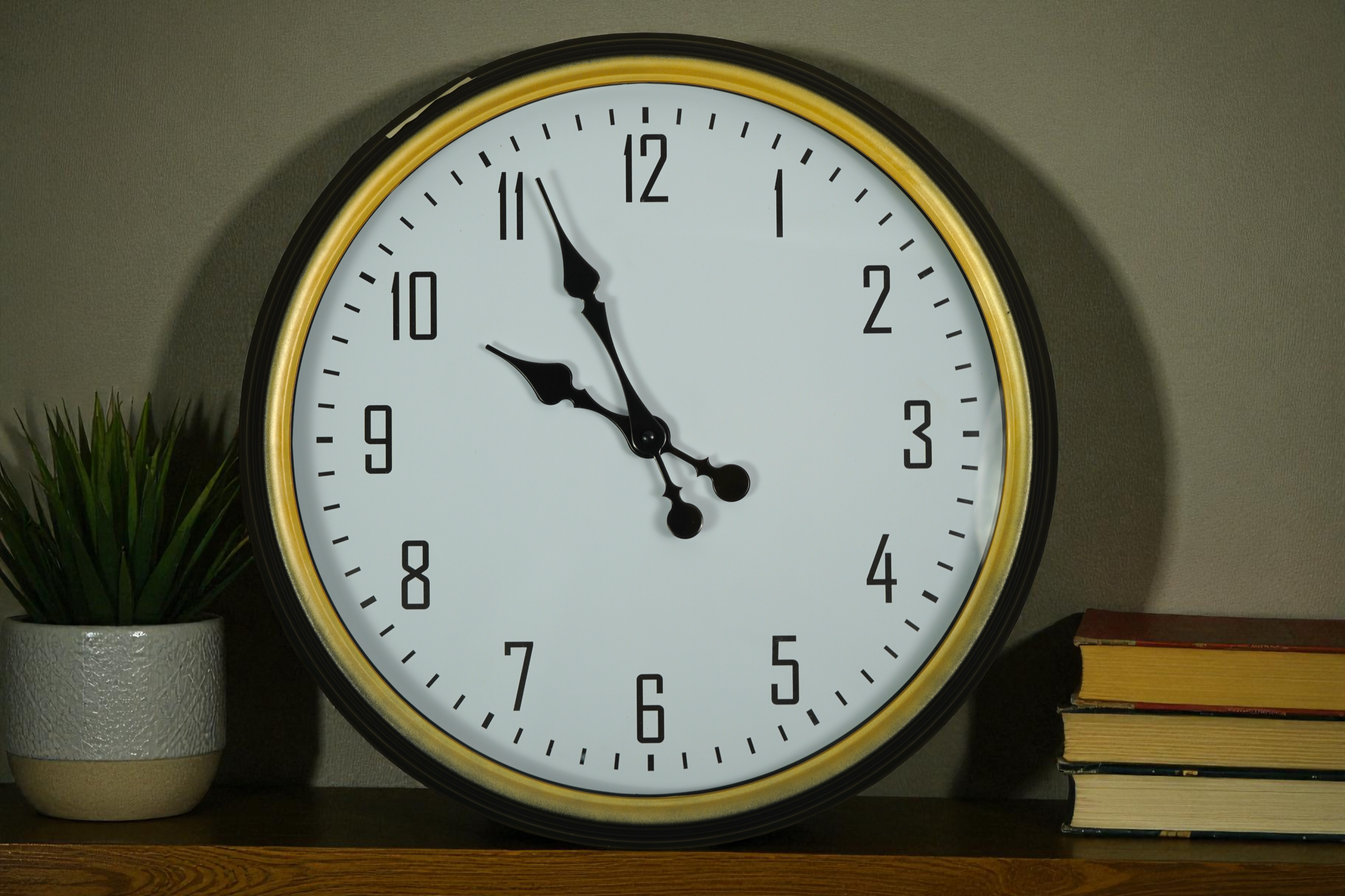 Black gold-rim classic wall clock on a shelf beside books and a plant