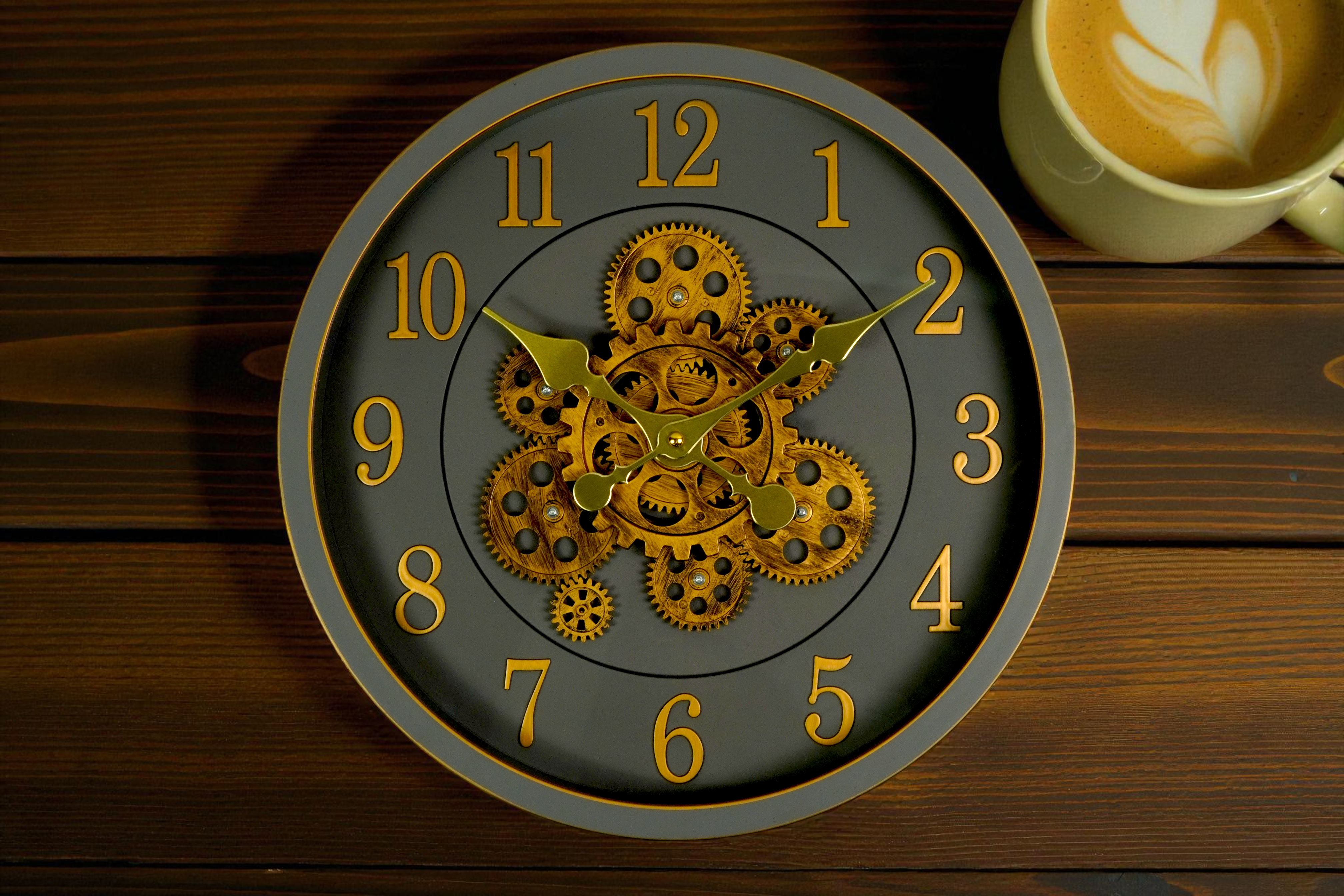 Grey gold gear skeleton wall clock on dark wood beside a coffee cup