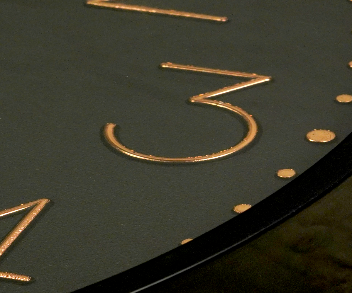 Matte black copper numeral wall clock on a grey surface in raking light