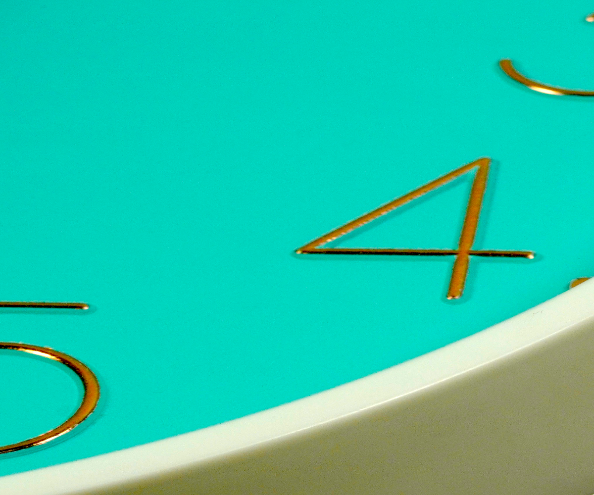 Turquoise gold numeral wall clock on teal planks beside a small plant